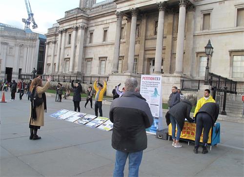 '图4：二零一九年二月二日，法轮功学员在伦敦特拉法加广场（Trafalgar Square）北平台的功法展示吸引过路民众现场学炼，图中一位西人女士正在模仿法轮功第二套功法“法轮桩法”中的“头顶抱轮”动作。'